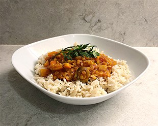 A White square bowl on a white marble background. the bowl is 1/2 full of brown basmati rice with a warm rich golden lentil chili over it and a sprig of mint on the top