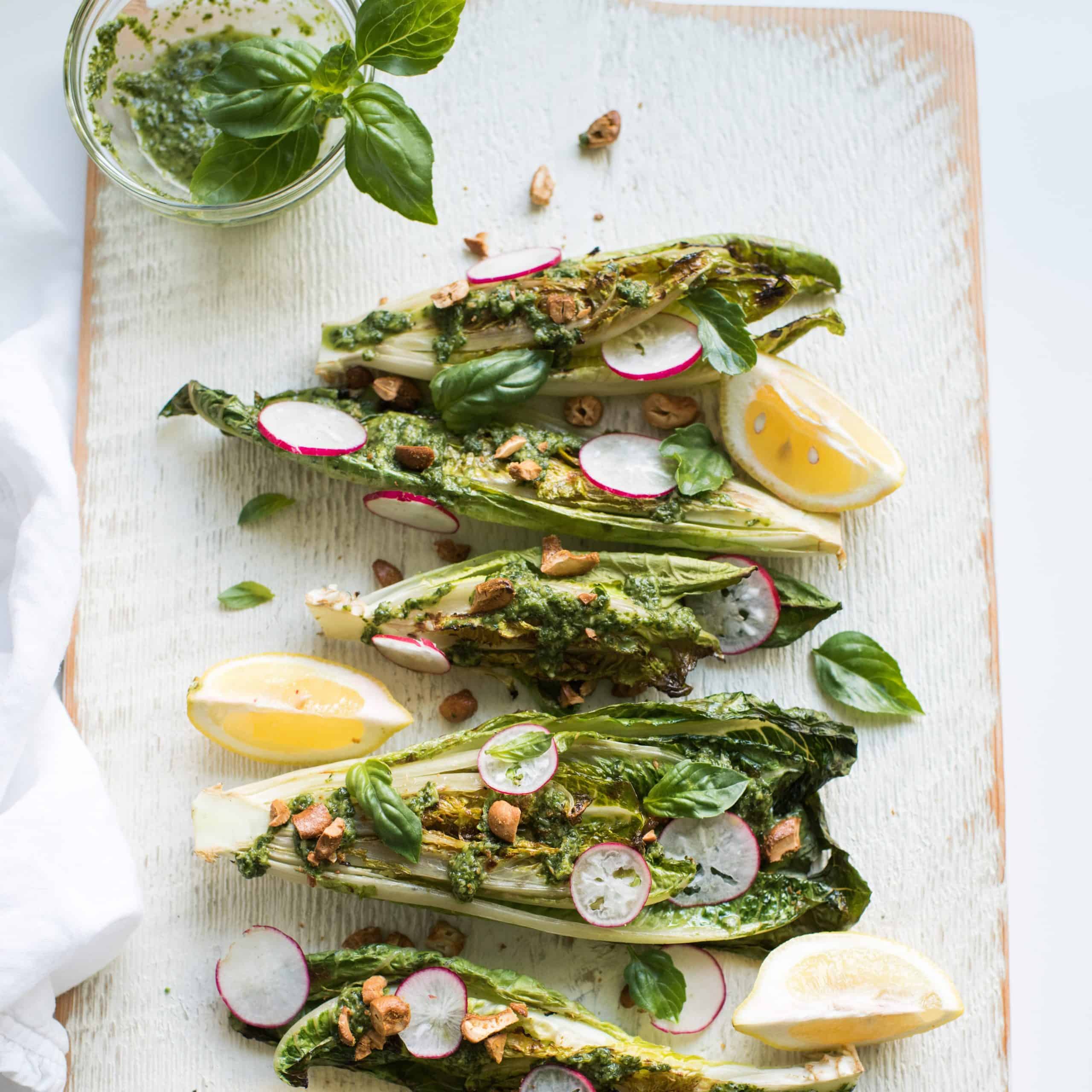 looking down on a cuting boad with 6 grilled hearts of romaine topped with slices radishes, basil leaves and chopped nuts. there are some lemon wedges here and thee too 