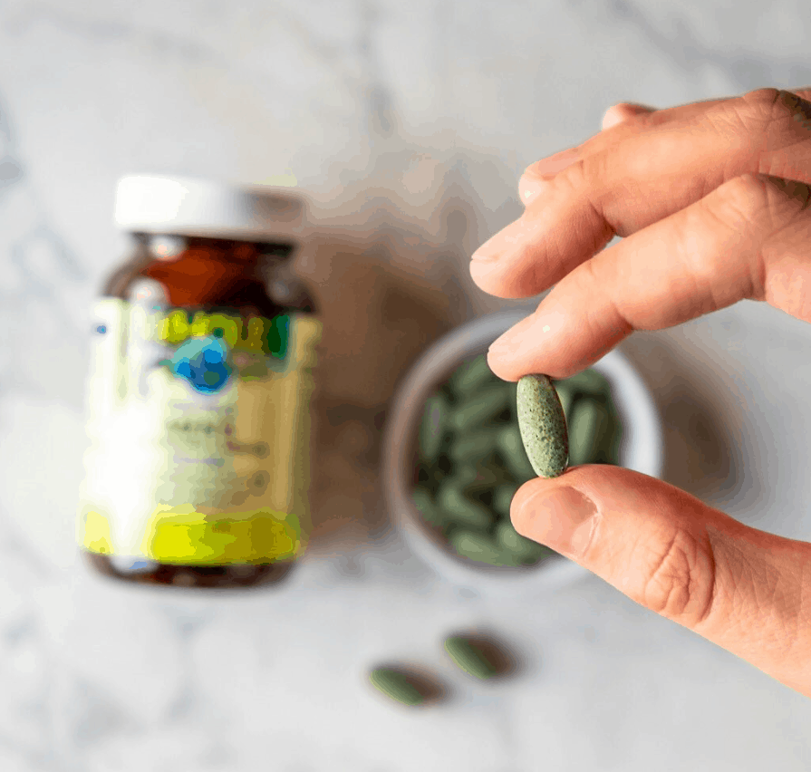 in the foreground is a hand holding a green supplement tablet beween the fingrs. in the background and blurred, is a small bowl of the supplement tablets and a bottle of WE&S Multis